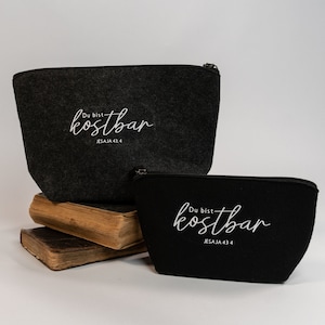 Könnte beinhalten: Zwei Kosmetiktaschen aus Filz mit Reißverschluss. Die größere Tasche ist grau und die kleinere ist schwarz. Beide Taschen haben weißen Text mit der Aufschrift "Du bist kostbar" und "Jesaja 43.4".