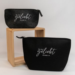 Könnte beinhalten: Zwei Reißverschlusstaschen, eine dunkelgraue und eine schwarze, mit dem deutschen Satz "Du bist geliebt" und dem Text "JOHANNES 3.16" in Weiß bedruckt. Die Taschen sind aus Filz.