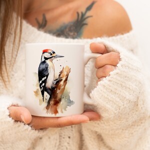 Puede incluir: Taza de cerámica blanca con una ilustración de acuarela de un pájaro carpintero posado en una rama. El pájaro tiene plumas blancas y negras y una cresta roja.