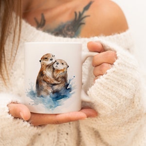 May include: White ceramic mug featuring a watercolour illustration of two otters embracing in the water. The otters are brown and have a light blue watercolour background.