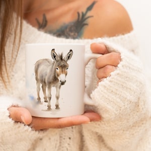 May include: A white ceramic mug featuring a watercolour illustration of a grey donkey. The mug is being held by a person wearing a white sweater.
