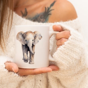 Puede incluir: Una taza de cerámica blanca con una ilustración de acuarela de un elefante bebé. El elefante es gris con una trompa y orejas de color marrón claro.