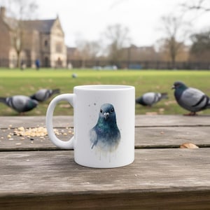 Pode incluir: Caneca de cerâmica branca com uma ilustração em aquarela de um pombo. A ave é retratada em tons de azul e cinza, com detalhes nas penas. A caneca está sobre uma mesa de madeira, com outros pombos ao fundo.