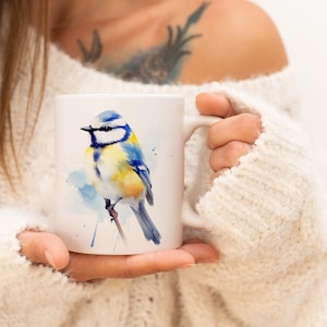 Op de afbeelding: Een witte keramische mok met een aquarel-illustratie van een blauwe mees op een tak. De vogel heeft blauwe, gele en witte veren. De mok wordt vastgehouden door een persoon die een witte trui draagt.