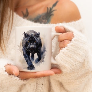 Könnte beinhalten: Eine weiße Keramiktasse mit einer Illustration einer schwarzen Pantherin. Die Pantherin ist in Aquarell-Stil mit einem wilden Ausdruck dargestellt.