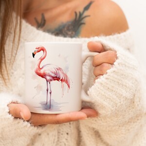 Puede incluir: Una taza de cerámica blanca con una ilustración de flamenco rosa. El flamenco está pintado en estilo acuarela con colores rosa, blanco y gris.