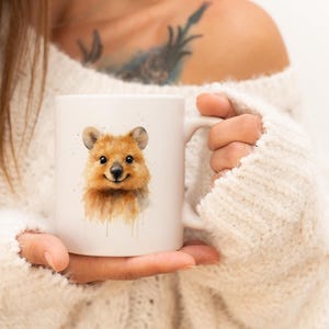 Può includere: Tazza in ceramica bianca con un'illustrazione ad acquerello di un quokka sorridente. Il quokka ha la pelliccia marrone e un'espressione amichevole. La tazza è tenuta da una persona che indossa un maglione bianco.
