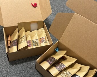 May include: Two open cardboard boxes filled with small, brown paper pouches. Each pouch has a clear window showing the contents, which appear to be dried herbs and flowers. The boxes are on a gray carpeted surface.
