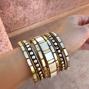 Può includere: Una collezione di braccialetti dorati con dettagli avorio e neri adorna un polso. I braccialetti presentano vari design, tra cui file di piccole perle dorate e intarsi rettangolari avorio, creando un aspetto stratificato e strutturato.