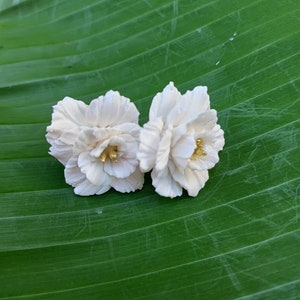 Puede incluir: Dos pendientes blancos en forma de flor con detalles dorados sobre un fondo de hoja verde.