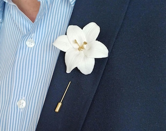 May include: A white lily flower lapel pin with gold accents, attached to a navy blue blazer. The pin is a stylish accessory. The background includes a light blue striped shirt.
