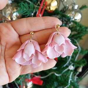 May include: A pair of pink flower-shaped dangle earrings with gold-tone posts. The earrings are made of polymer clay and have a layered, ruffled design.
