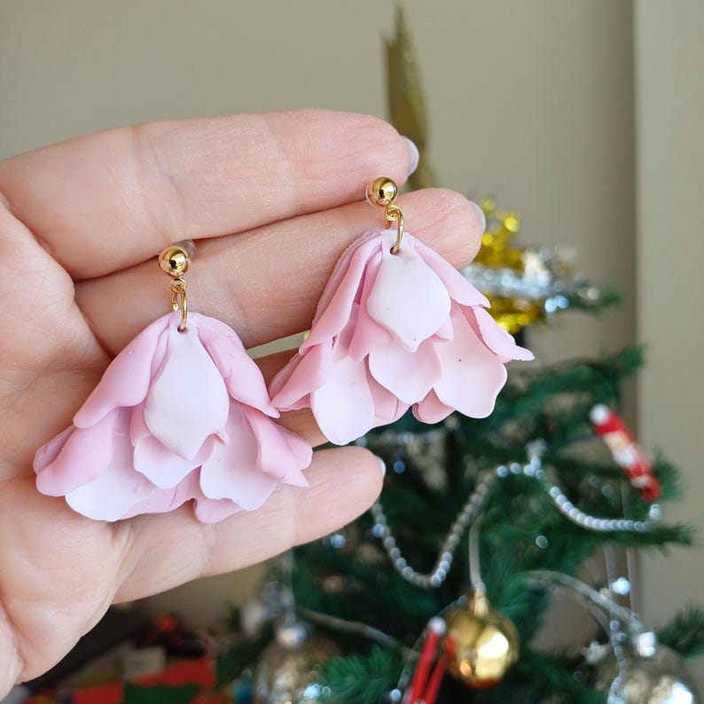 May include: A pair of pink and white flower-shaped dangle earrings with gold posts. The earrings are made of polymer clay and have a layered design.