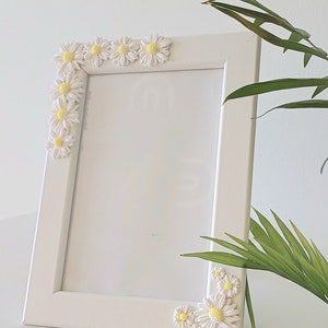 May include: White picture frame with a white border and a white mat. The frame is decorated with small white daisies with yellow centers around the edges.