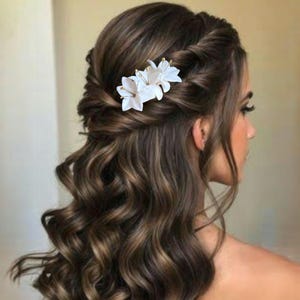 以下が含まれることがあります： 白い花の形をしたヘアクリップで、金色のアクセントが付いています。クリップは女性の茶色の髪に付いています。