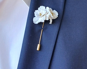 May include: A white flower lapel pin with gold accents is pinned to a navy blue blazer. The pin features three delicate flower blooms and a gold-colored pin. The blazer is paired with a white dress shirt.
