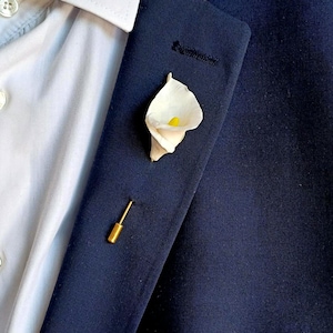 Könnte beinhalten: Ein weißer Calla-Lily-Anstecker mit gelber Mitte, an einem dunkelblauen Sakko befestigt.