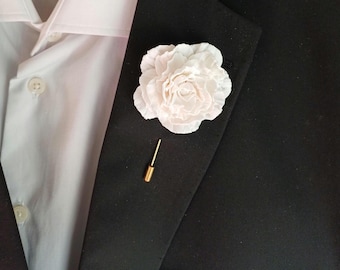 May include: A white fabric flower boutonniere with a gold pin. The flower is on a black suit jacket.