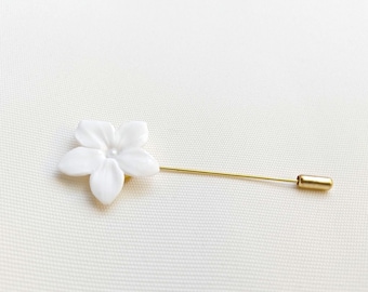 May include: A white flower lapel pin with a gold-colored pin. The flower has five petals and a small pearl-like center. The pin is a simple, elegant accessory.