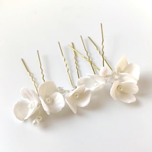 May include: A set of white floral hair pins with gold-coloured stems and small pearl accents. The delicate flower petals are arranged on the pins, creating an elegant look. Ideal for weddings or special occasions.