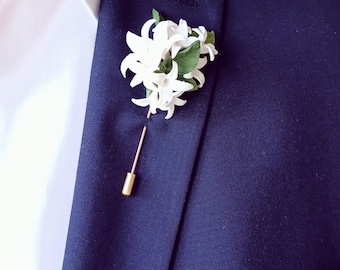 Puede incluir: Un prendedor floral blanco con hojas verdes y un alfiler dorado está sujeto a una blazer azul marino. El prendedor es un accesorio decorativo, a menudo utilizado para eventos formales o para añadir elegancia.