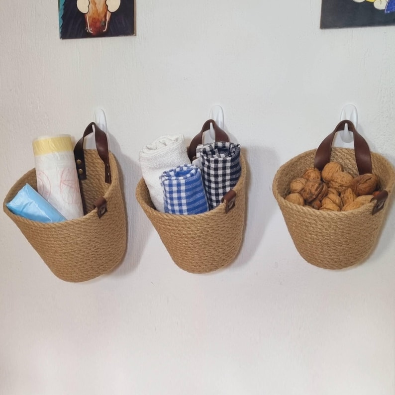 May include: Three brown rope wall baskets with leather handles. The baskets are hanging on white walls and contain various items, including a blue and white checkered dish towel, a white dish towel, and a pile of walnuts.