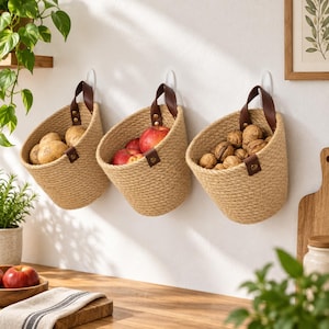 Op de afbeelding: Drie aan de muur gemonteerde opbergmanden van gevlochten beige jutekoord, elk voorzien van een donkerbruine leren riem die is vastgezet met metalen klinknagels. De manden zijn gevuld met respectievelijk aardappelen, rode appels en walnoten.