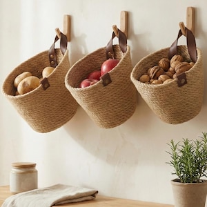 May include: Three woven jute baskets with leather handles hang on a wall, each filled with produce. One contains potatoes, another red apples, and the third walnuts. The baskets are suspended from wooden pegs, providing a rustic storage solution for the kitchen.