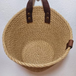 May include: A round, tan, woven jute basket with brown leather handles and a brown leather strap on the side. The basket is empty.