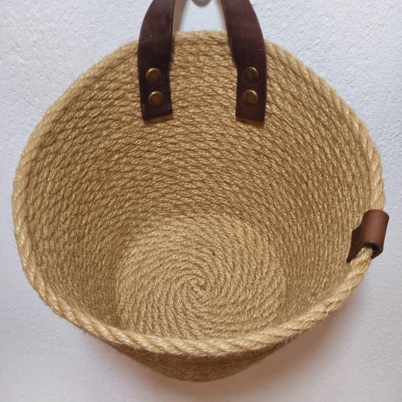 May include: A round, tan, woven jute basket with brown leather handles and a brown leather strap on the side. The basket is empty.