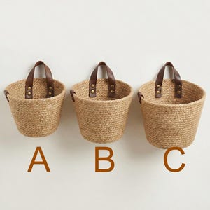 May include: Three woven jute baskets with brown leather handles and brass rivets are displayed against a white wall. The baskets are labeled A, B, and C in brown capital letters. The baskets are a natural tan color.