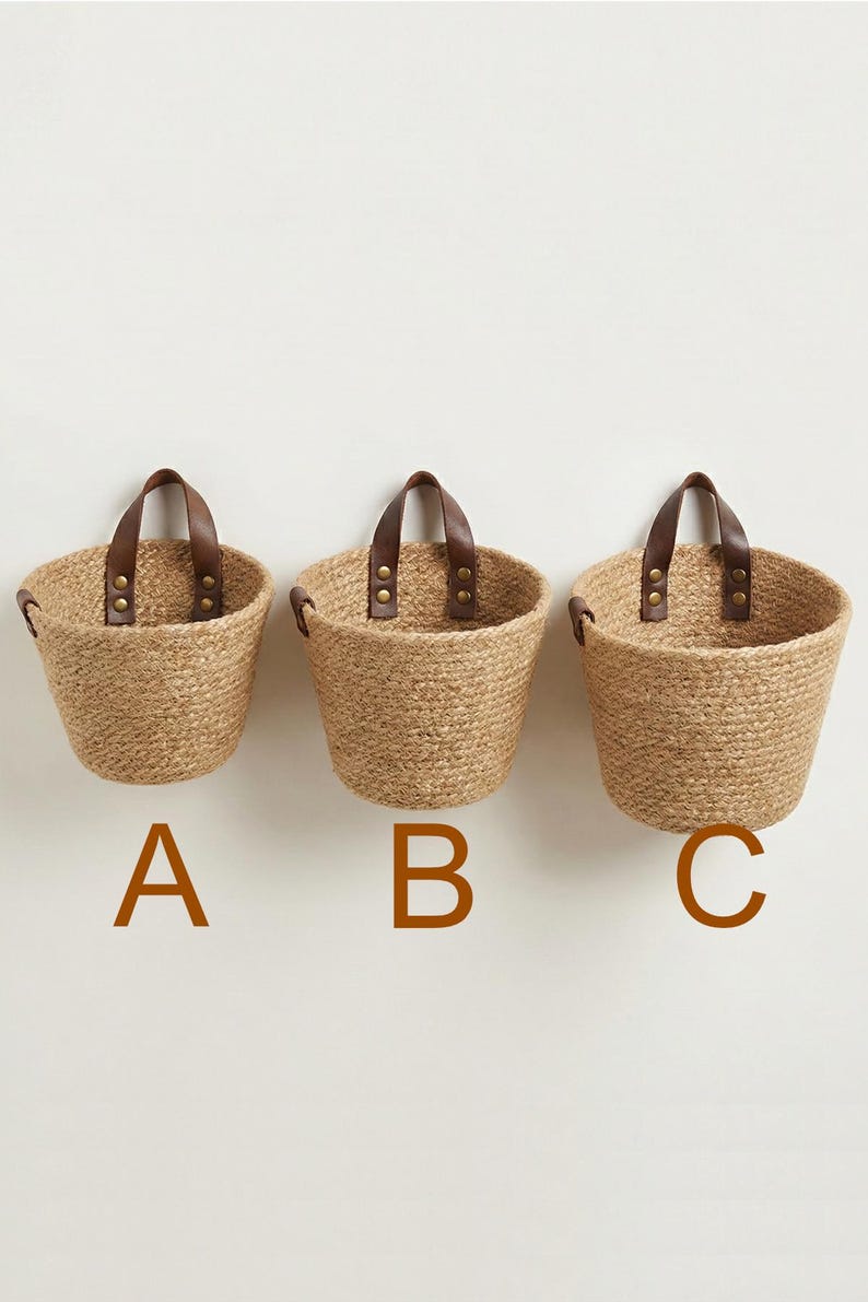 May include: Three woven jute baskets with brown leather handles and brass rivets are displayed against a white wall. The baskets are labeled A, B, and C in brown capital letters. The baskets are a natural tan color.