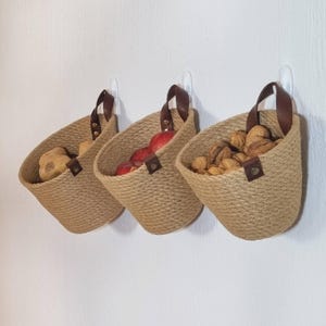 May include: Three wall-mounted jute baskets with leather handles. The baskets are filled with potatoes, apples, and walnuts.