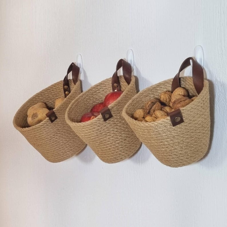 May include: Three wall-mounted jute baskets with leather handles. The baskets are filled with potatoes, apples, and walnuts.