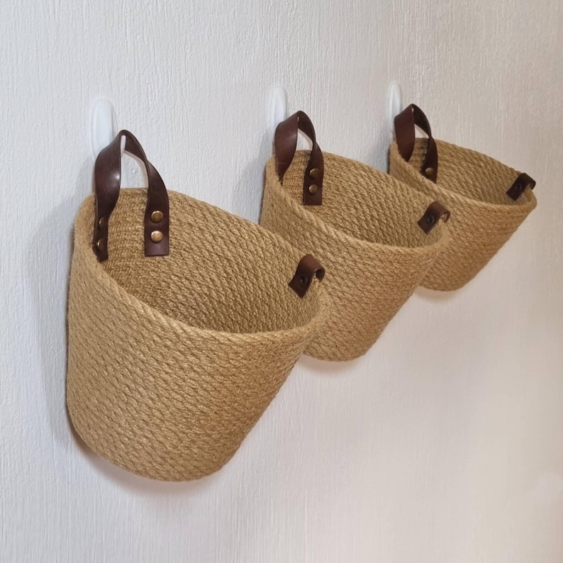 May include: Three brown rope wall baskets with leather handles and brass accents. The baskets are hanging on white hooks against a white wall.