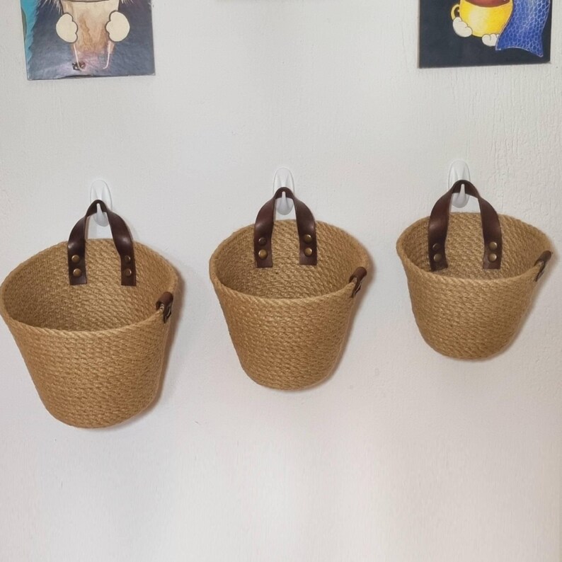 May include: Three brown woven wall baskets with leather handles and brass accents. The baskets are hanging on a white wall.
