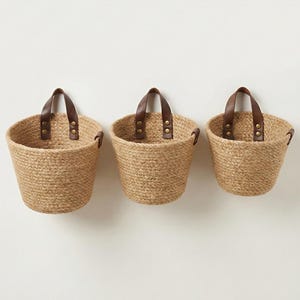 May include: Three woven jute baskets with brown leather handles attached with brass rivets. The baskets are of varying sizes and are designed to be hung on a wall. The natural color of the jute provides a rustic aesthetic.