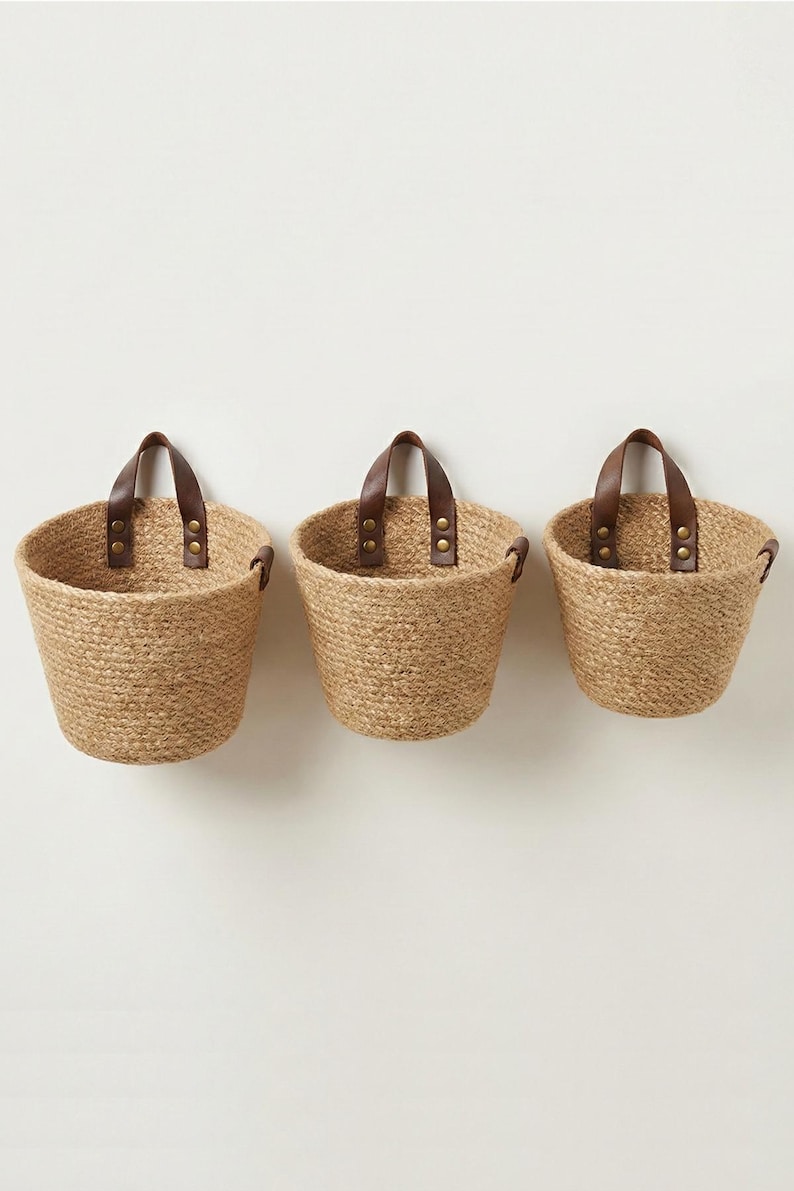 May include: Three woven jute baskets with brown leather handles attached with brass rivets. The baskets are of varying sizes and are designed to be hung on a wall. The natural color of the jute provides a rustic aesthetic.