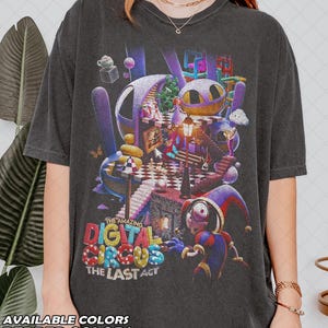 May include: A charcoal gray t-shirt featuring a vibrant, multi-colored graphic print of The Amazing Digital Circus The Last Act. The design includes a jester character, a checkered floor, a street lamp, and various surreal, colorful digital elements.
