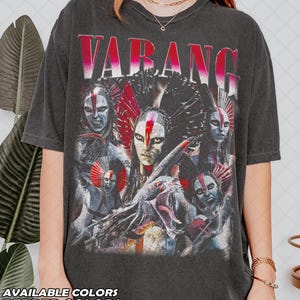 Pode incluir: T-shirt cinzenta escura com um gráfico com a palavra "VARANG" em vermelho e branco, e uma ilustração detalhada de várias figuras com pintura facial tribal e cocares. O texto "AVAILABLE COLORS" e "PEPPER COLORS" está na parte inferior.