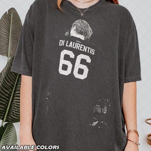 May include: A charcoal gray short-sleeve t-shirt featuring a white graphic print on the chest. The design includes the text DI LAURENTIS above the number 66, accompanied by a stylized illustration of a person viewed from behind.