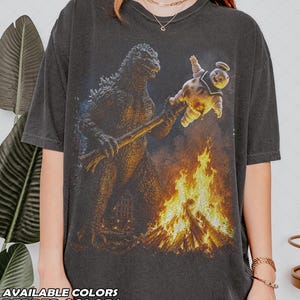 May include: A charcoal gray t-shirt featuring a graphic print of Godzilla holding a stick with a Stay Puft Marshmallow Man over a campfire. The design includes vibrant orange and yellow flames, with the text AVAILABLE COLORS PEPPER COLORS.