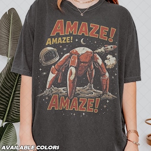 May include: A charcoal gray t-shirt featuring a graphic of a red robotic creature on a moon surface, an astronaut helmet, a rocket, and stars. The text AMAZE! appears in red at the top and bottom. The design has a distressed, vintage-inspired aesthetic.