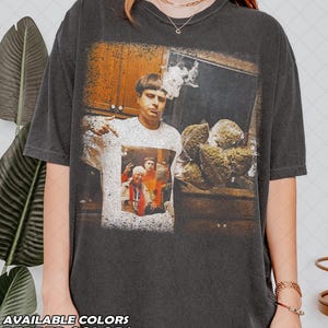 May include: A charcoal gray t-shirt featuring a graphic print of a person with a bowl cut hairstyle. The graphic shows the person in a kitchen setting, holding a large cluster of green plant buds, with a smaller inset image of the same person.