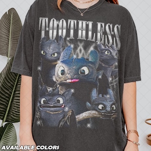 May include: Dark grey t-shirt featuring a graphic of Toothless from How to Train Your Dragon. The design includes multiple images of Toothless with the word "TOOTHLESS" in silver text. The bottom of the shirt has the text "AVAILABLE COLORS PEPPER COLORS".
