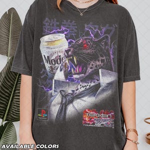 Puede incluir: Camiseta gris oscuro con un diseño de jaguar con ojos rojos, una lata de cerveza y texto japonés. Incluye los logotipos de PlayStation y Tekken Tag Tournament. Disponible en colores pimienta.