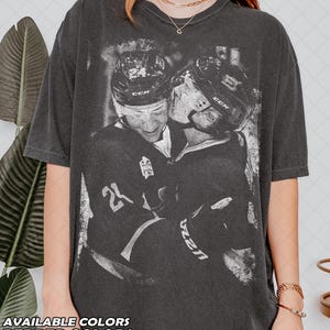 May include: Dark grey t-shirt with a black and white image of two hockey players in helmets, hugging. The shirt displays the number 24. The text "AVAILABLE COLORS" and "PEPPER COLORS" is at the bottom of the shirt.