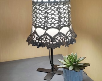 May include: A crocheted lampshade in a light beige color with a delicate pattern. The lampshade is on a black metal lamp base with a cross-shaped stand. A small succulent plant in a white ceramic pot is on the table next to the lamp.