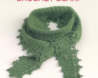 Può includere: Una sciarpa verde all'uncinetto con un delicato bordo in pizzo. La sciarpa è piegata a metà e ha un piccolo ciondolo a forma di cuore.