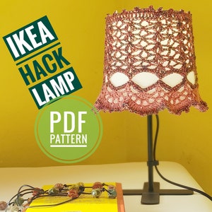 May include: A crocheted lampshade in shades of pink and brown with a black metal lamp base. The lampshade has a scalloped edge and a delicate floral pattern. The image includes the text "IKEA HACK LAMP PDF PATTERN".